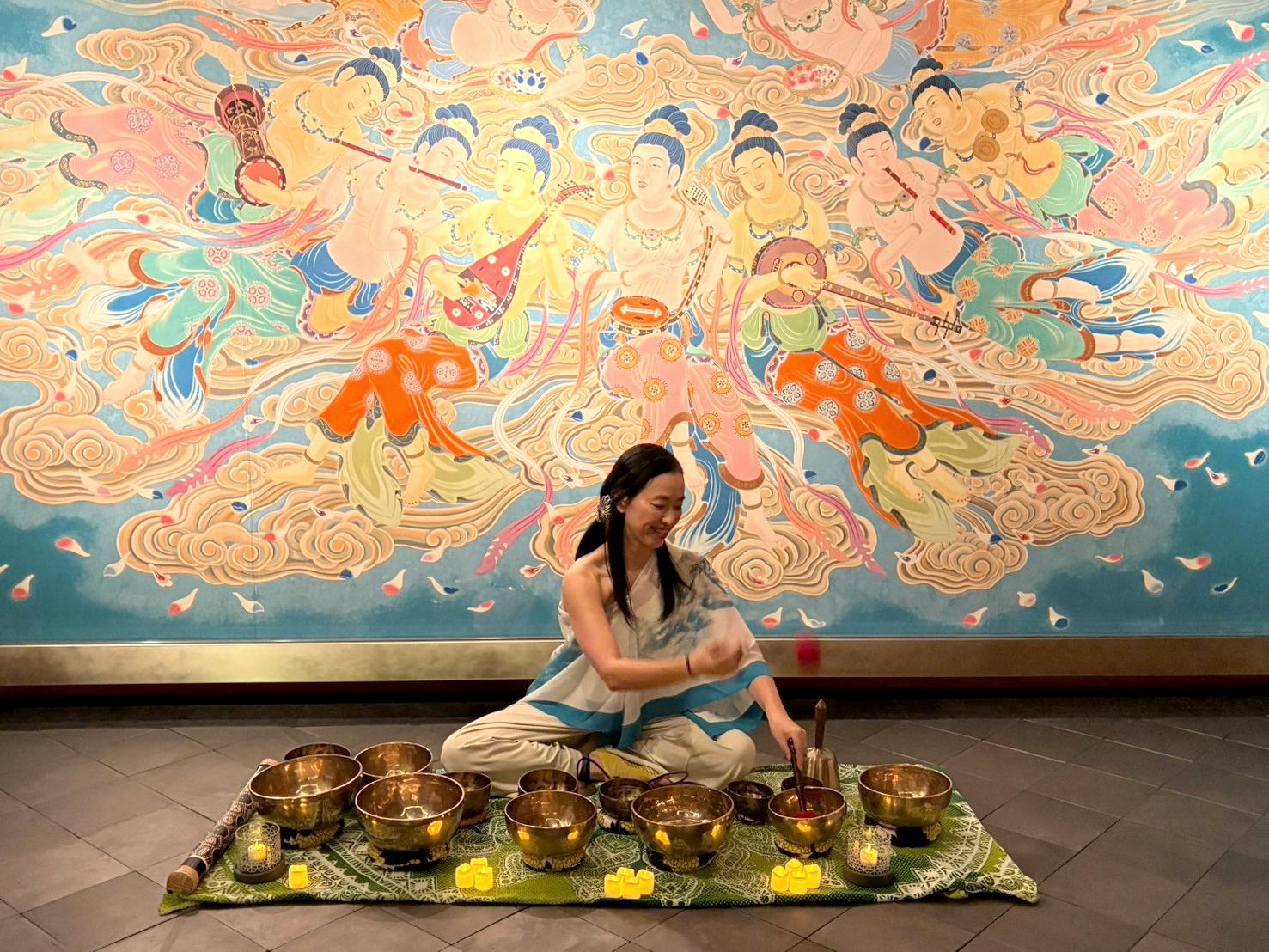Singing bowls of several sizes on a green cloth in front of the mural, with the player reaching toward one with a mallet.