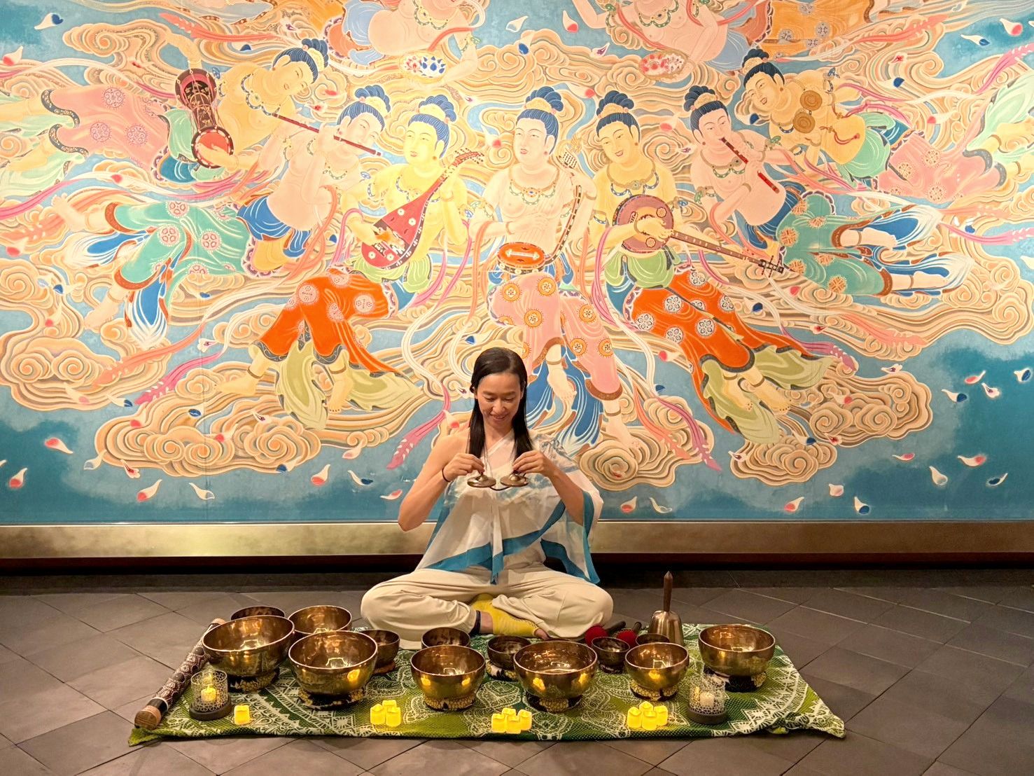 A woman sits cross-legged in front of a large painted mural, holding a pair of small hand cymbals, with singing bowls and small candle lights laid out before her.
