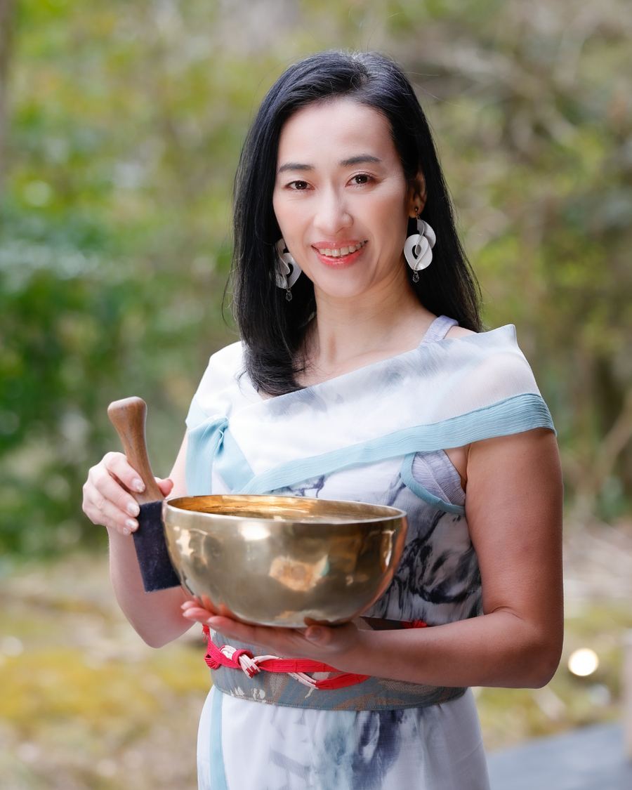 Yasuko Kato holding a singing bowl and mallet, in a green garden.