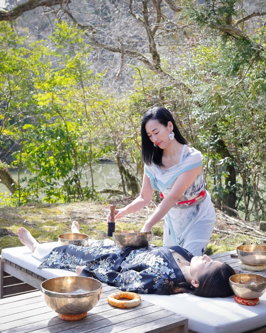 A person lies resting with eyes closed while a singing bowl is played beside them.